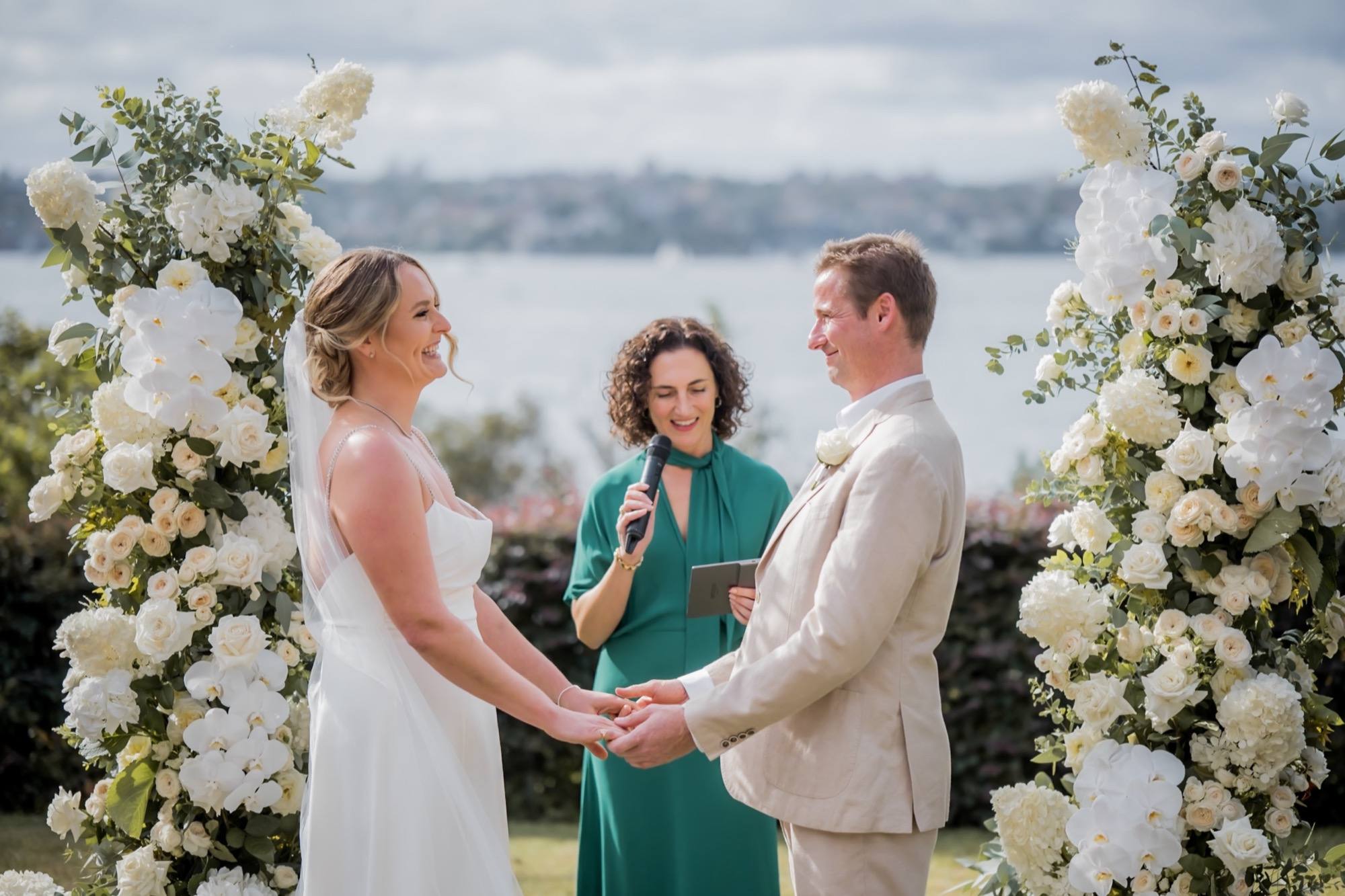 A couple exchanging vows in front of white floral arches overlooking Sydney Harbour