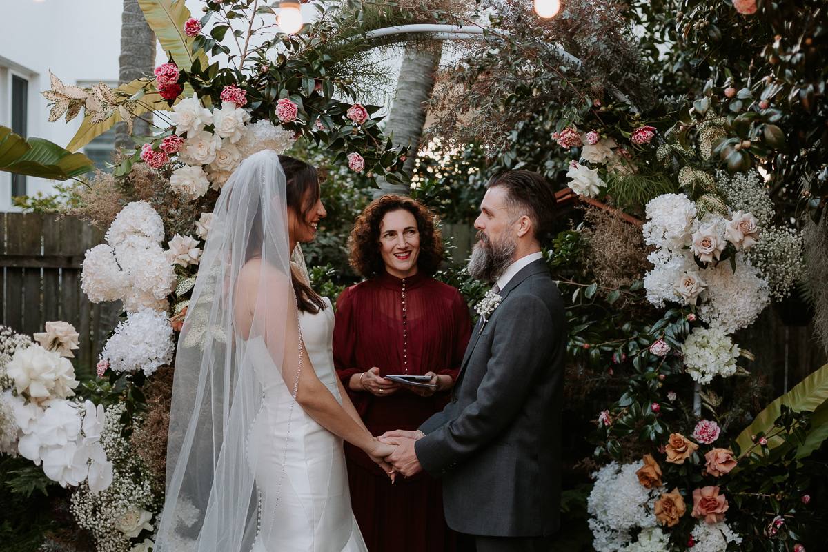 A couple holding hands beneath a lush floral arch during an evening garden ceremony