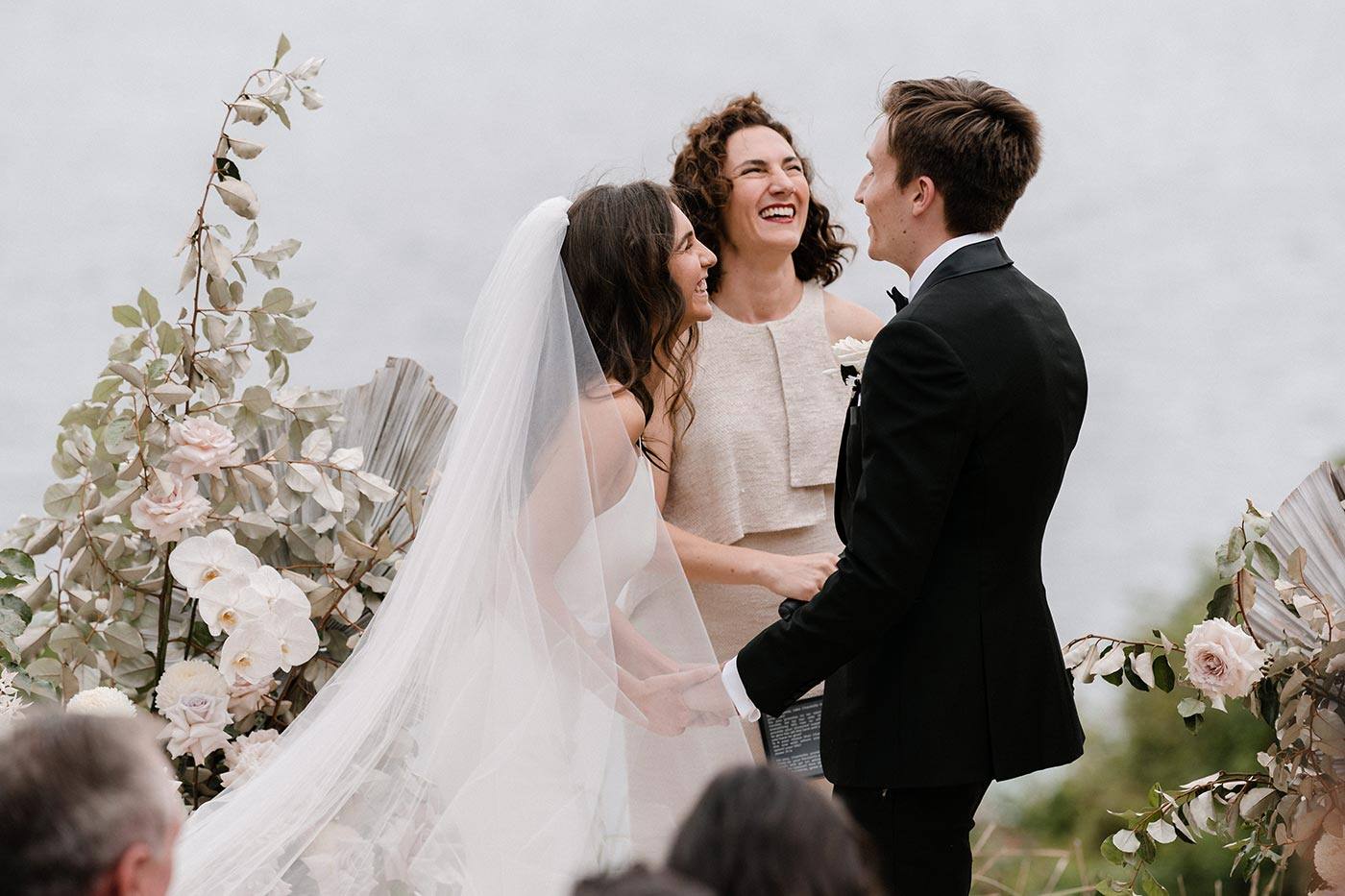 A bride and groom laughing with their marriage celebrant during a waterfront wedding ceremony